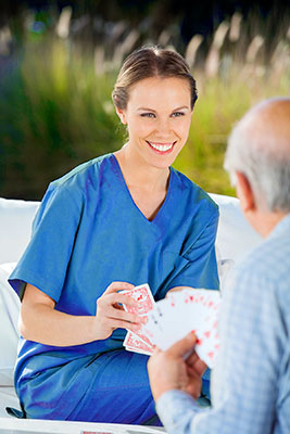 beautiful whil female caregiver in bright blue scrubs smiling and playing cards with an elderly man in a long-sleeved blue shirt