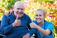 elderly white man sitting with a young pretty white female caregiver in blue scrubs with both giving a thumbs up