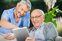 happy male caregiver in blue scrubs and smiling white senior man looking at ipad